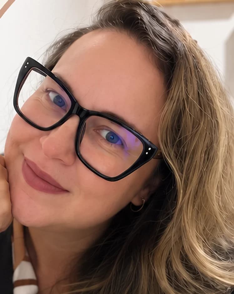 Karina Coutinho, psicóloga, sorrindo, de óculos de armação preta, com o rosto apoiado na mão